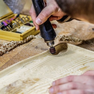 May include: A person uses a rotary tool with a sanding attachment to smooth a wooden surface. The tool is black and has a brown sanding disc. The wood is light brown and has a grain pattern. There are wood shavings on the surface.