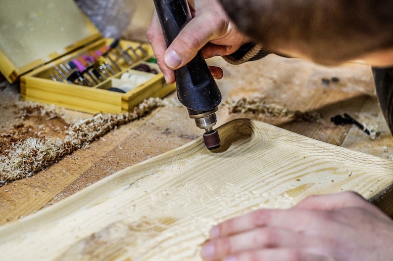 May include: A person uses a rotary tool with a sanding attachment to smooth a wooden surface. The tool is black and has a brown sanding disc. The wood is light brown and has a grain pattern. There are wood shavings on the surface.