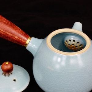 Può includere: Teiera in ceramica azzurra con manico e coperchio in legno. Il manico è marrone rossiccio con una fascia dorata. Il coperchio ha un piccolo pomello in legno con base metallica decorativa. Include un filtro.