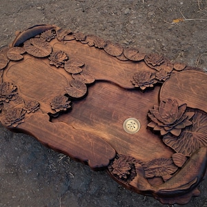 May include: A wooden serving tray carved with a lotus pond design. The tray features intricate details of lotus flowers, leaves, and fish. A small, round, brass plate with the Chinese character for "tea" is centered on the tray.