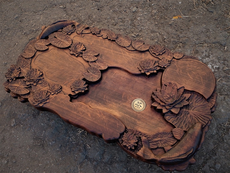May include: A wooden serving tray carved with a lotus pond design. The tray features intricate details of lotus flowers, leaves, and fish. A small, round, brass plate with the Chinese character for "tea" is centered on the tray.