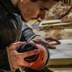 May include: A person uses a black and orange electric sander to smooth a piece of wood.