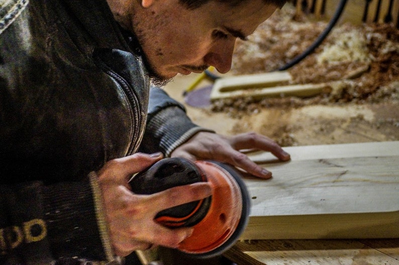 May include: A person uses a black and orange electric sander to smooth a piece of wood.