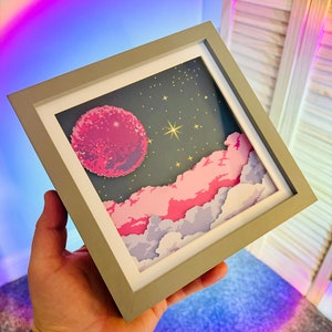 Op de afbeelding: Een ingelijst pixel art-print met een roze planeet met een ster in een donkerblauwe lucht met roze wolken. De lijst is grijs met een witte passe-partout.