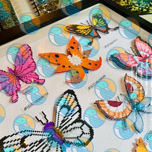 May include: A framed display featuring multiple pixel art butterfly figures crafted from small plastic beads. The collection includes labeled specimens such as #0049 VENOMOTH, #0637 VOLCARONA, #0267 BEAUTIFLY, and #0012 BUTTERFREE, arranged on a white background.