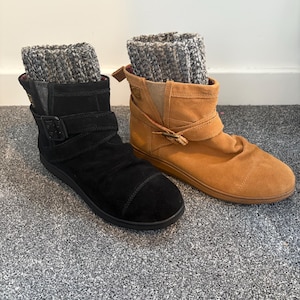 May include: A pair of ankle boots, one black and one tan, both featuring a rounded toe, a side buckle strap, and a thick, grey, ribbed knit cuff extending from the top. The boots have a flat sole and a soft, suede-like exterior texture.