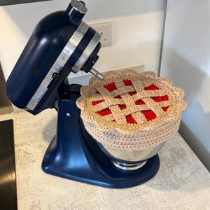 May include: A dark blue KitchenAid Artisan stand mixer with a stainless steel bowl covered by a round, crocheted pie-shaped lid. The lid features a tan lattice crust design over a bright red filling, with a scalloped edge and a textured finish.