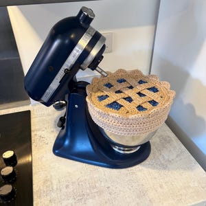 May include: A dark blue KitchenAid Artisan stand mixer with a stainless steel bowl covered by a crocheted pie-shaped topper. The topper features a tan lattice crust design over a dark blue filling, with a scalloped edge around the circumference.