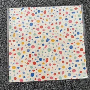 May include: A square paper napkin featuring a cream-coloured background with a scattered pattern of small, multi-coloured dots in shades of red, blue, yellow, and light green. The napkin is enclosed in a clear, protective plastic sleeve.