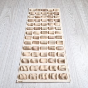 Puede incluir: Una esterilla de masaje de pies beige con numerosos bloques de madera texturizados y elevados dispuestos en filas. La esterilla es rectangular y está diseñada para reflexología podal. Los bloques de madera son de color marrón claro.