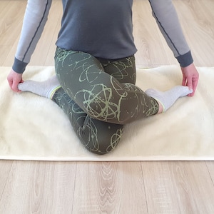 May include: A person is sitting on a white yoga mat with their legs crossed. They are wearing gray and green leggings with a green and white abstract pattern. They are also wearing white socks.