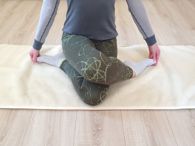 May include: A person is sitting on a white yoga mat with their legs crossed. They are wearing gray and green leggings with a green and white abstract pattern. They are also wearing white socks.