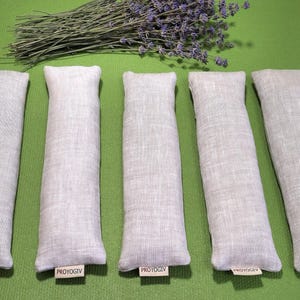 May include: Five rectangular, pale grey linen sachets with rounded tops are arranged on a green surface. A bundle of dried lavender with purple flowers rests above them. Each sachet has a small tag that reads "PROYOGIV".