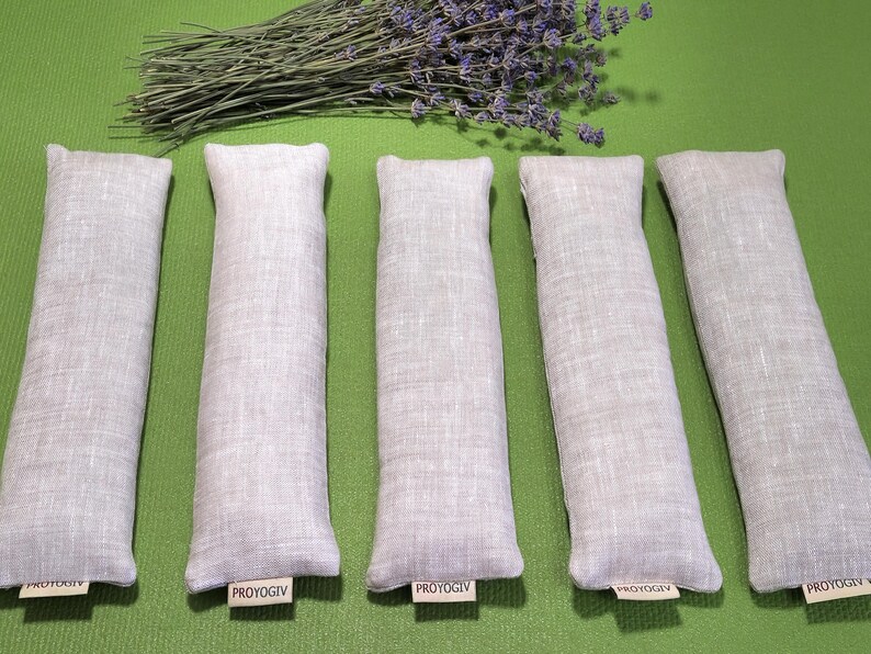 May include: Five rectangular, pale grey linen sachets with rounded tops are arranged on a green surface. A bundle of dried lavender with purple flowers rests above them. Each sachet has a small tag that reads "PROYOGIV".
