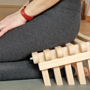 Könnte beinhalten: Eine hellfarbene Meditationsbank aus Holz mit einer Lattenrost-Sitzfläche und abgewinkelten Beinen. Die Bank ist so konzipiert, dass sie die Körperhaltung des Benutzers während der Meditation unterstützt.
