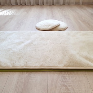 May include: A white faux fur rug with a green backing. The rug is laid out on a light wood floor. Two white round cushions are in the background.