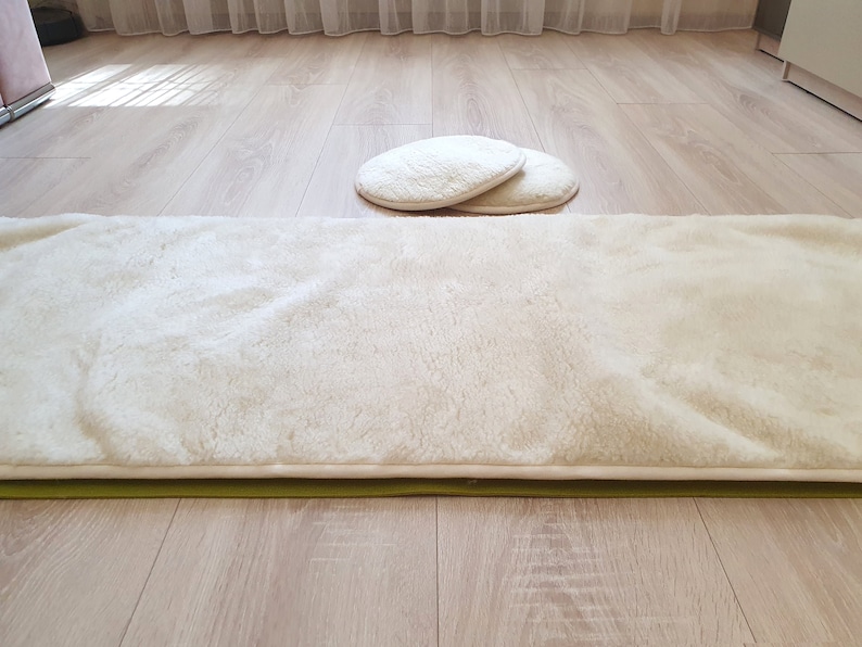 May include: A white faux fur rug with a green backing. The rug is laid out on a light wood floor. Two white round cushions are in the background.