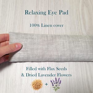 May include: A light grey linen eye pad with the text "Relaxing Eye Pad" and "100% Linen cover". The eye pad is filled with flax seeds and dried lavender flowers. The eye pad is approximately 20 cm long.
