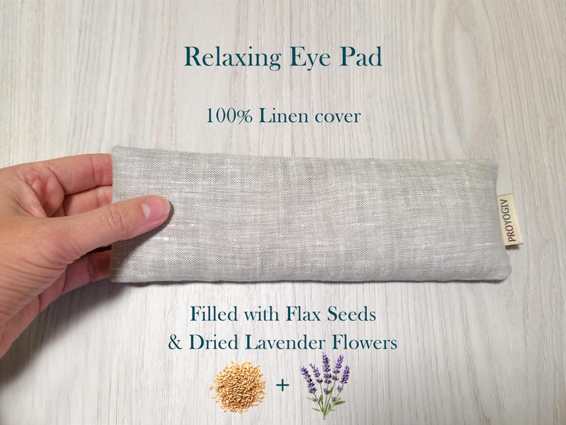 May include: A light grey linen eye pad with the text "Relaxing Eye Pad" and "100% Linen cover". The eye pad is filled with flax seeds and dried lavender flowers. The eye pad is approximately 20 cm long.