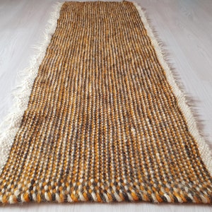 May include: A long, narrow, woven rug with a fringe border. The rug is made of a mix of brown, white, and orange yarn.