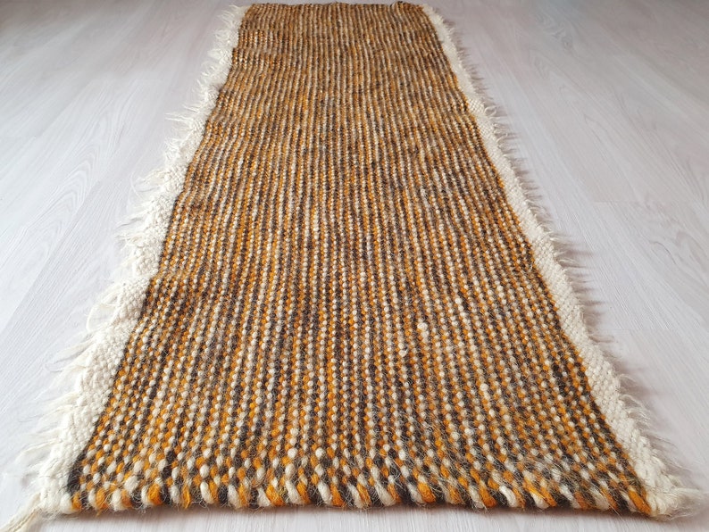 May include: A long, narrow, woven rug with a fringe border. The rug is made of a mix of brown, white, and orange yarn.