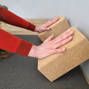 May include: Two brown cork yoga blocks are stacked on top of each other. A person's hands are resting on the top block.