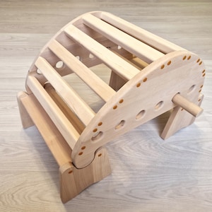 May include: A natural wood climbing arch with a curved ladder design. The wooden structure features multiple rungs and a rounded top, supported by two angled legs. Small, decorative pegs are visible along the edges.