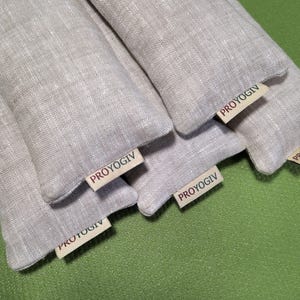 May include: Several light grey linen pouches are displayed, each with a small tag bearing the text "PROYOGIV". The pouches are arranged on a green surface. The tags are visible on the front of each pouch. The pouches appear to be filled.