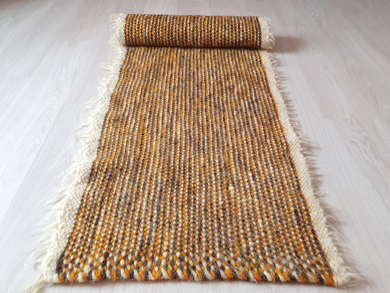May include: A long, narrow, handwoven rug with a fringe on both ends. The rug is made of wool in shades of brown, orange, and white.