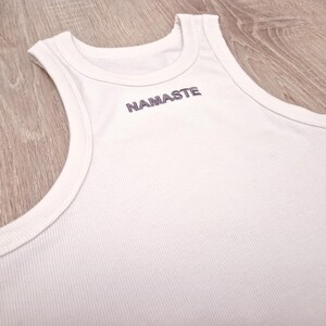 K&ouml;nnte beinhalten: Wei&szlig;es geripptes Tanktop mit dem Wort "NAMASTE" auf der Vorderseite gestickt.
