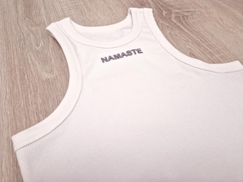 K&ouml;nnte beinhalten: Wei&szlig;es geripptes Tanktop mit dem Wort "NAMASTE" auf der Vorderseite gestickt.