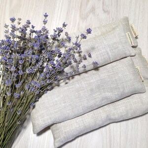 May include: A bunch of dried lavender with purple flowers beside three rectangular, light grey linen sachets. Each sachet has a small, rectangular tag with the text "AIDOAIOA" printed on it. The items are arranged on a light wood surface.