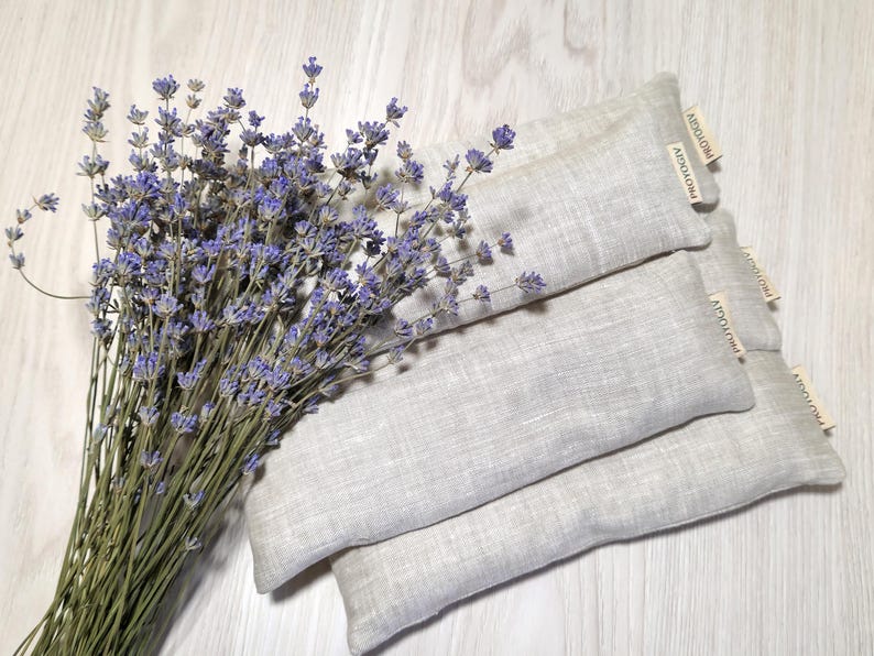 May include: A bunch of dried lavender with purple flowers beside three rectangular, light grey linen sachets. Each sachet has a small, rectangular tag with the text "AIDOAIOA" printed on it. The items are arranged on a light wood surface.