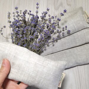 May include: A bunch of dried lavender with purple flowers and several grey linen aromatherapy pillows. One pillow is held in a hand. The pillows have a rectangular shape and a tag that reads "PROYOGIV".