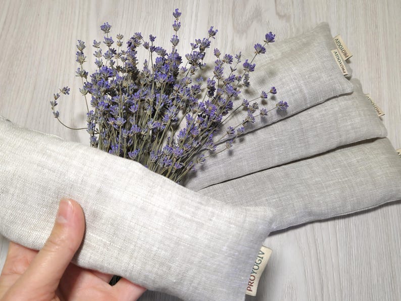 May include: A bunch of dried lavender with purple flowers and several grey linen aromatherapy pillows. One pillow is held in a hand. The pillows have a rectangular shape and a tag that reads "PROYOGIV".