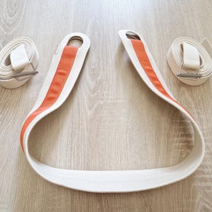 May include: A white and orange strap with two metal rings. The strap is made of a thick, woven material and has a bright orange stripe down the centre.