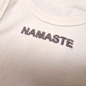 K&ouml;nnte beinhalten: Wei&szlig;es Tanktop mit dem Wort "NAMASTE" in Grau auf der Vorderseite gestickt.