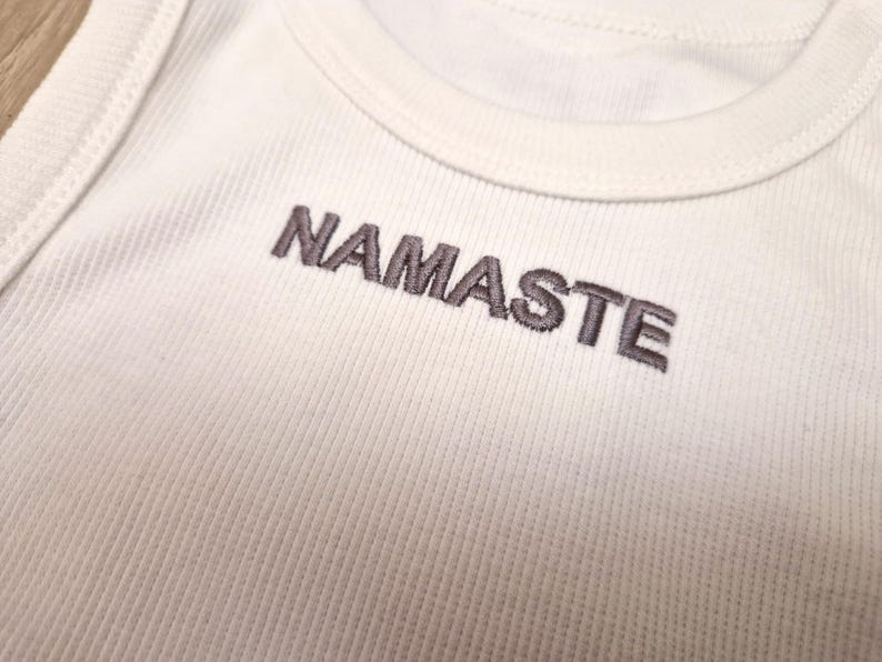 K&ouml;nnte beinhalten: Wei&szlig;es Tanktop mit dem Wort "NAMASTE" in Grau auf der Vorderseite gestickt.