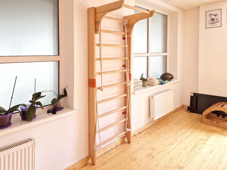 May include: Wooden wall-mounted Swedish ladder with horizontal rungs and a rope. The ladder is made of light-colored wood and is attached to a white wall. The ladder is designed for exercise and physical therapy.