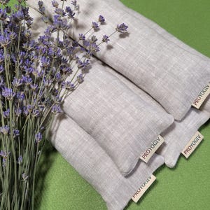 May include: A bunch of dried lavender next to several light grey linen aromatherapy pillows. Each pillow has a small tag that reads "PROYOGIV". The pillows are arranged on a green surface.