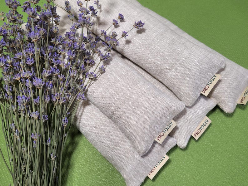 May include: A bunch of dried lavender next to several light grey linen aromatherapy pillows. Each pillow has a small tag that reads "PROYOGIV". The pillows are arranged on a green surface.