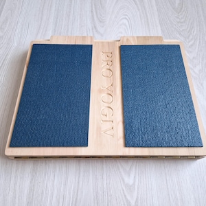 Op de afbeelding: Een houten voetensteun met twee marineblauwe, getextureerde pads. De tekst "PRO YOGIV" is in het hout tussen de pads gegraveerd. De voetensteun is ontworpen voor yoga of andere oefeningen.