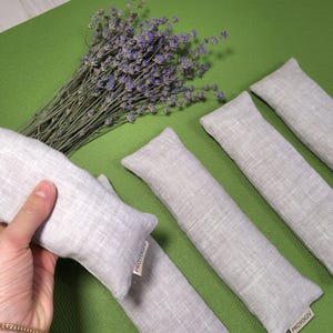 May include: A collection of light grey linen aromatherapy pillows, alongside a bunch of dried lavender, are displayed on a green surface. One pillow is held by a hand. Each pillow has a small tag with text.