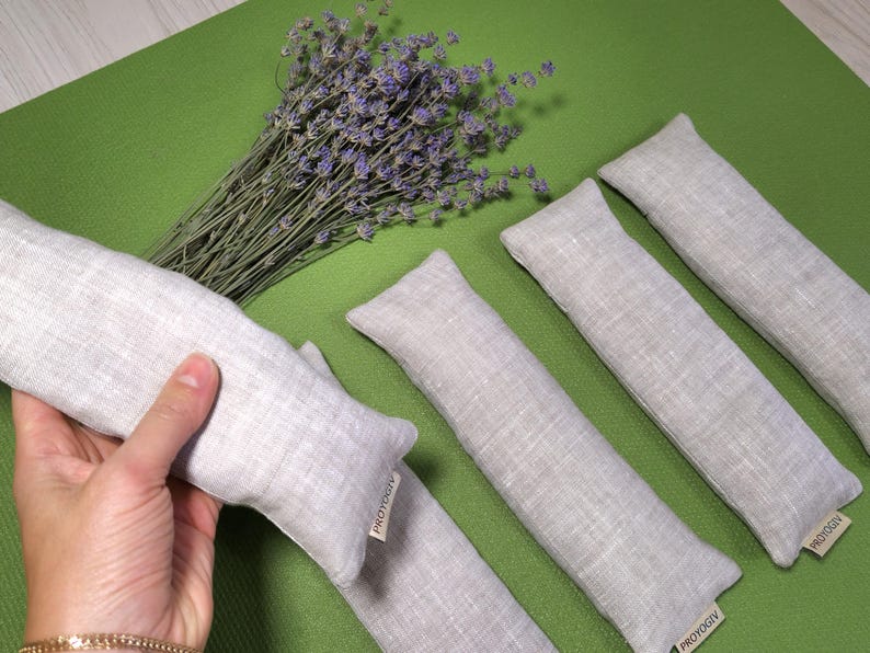 May include: A collection of light grey linen aromatherapy pillows, alongside a bunch of dried lavender, are displayed on a green surface. One pillow is held by a hand. Each pillow has a small tag with text.