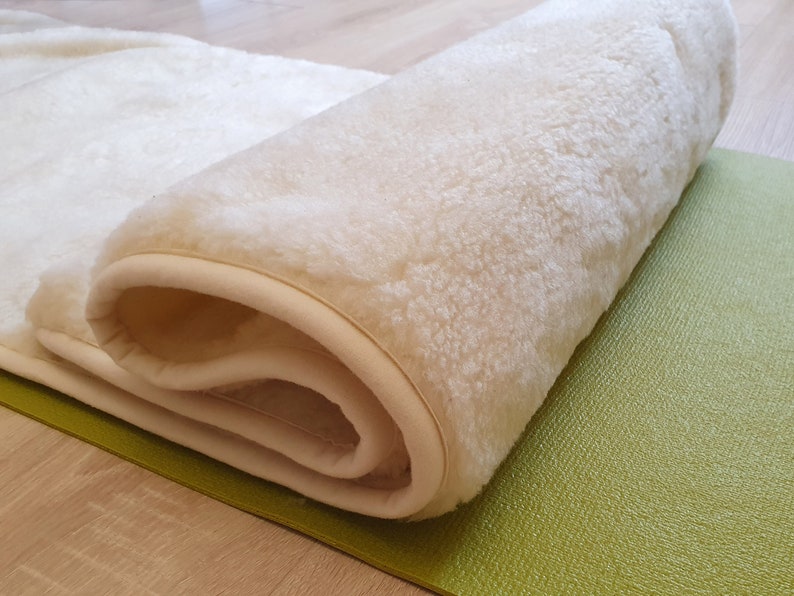 May include: A white sheepskin yoga mat rolled up on a green yoga mat. The sheepskin mat has a cream colored border.