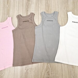 K&ouml;nnte beinhalten: Vier &auml;rmellose Tanktops in verschiedenen Farben: rosa, braun, grau und wei&szlig;. Jedes Tanktop hat den Schriftzug "NAMASTE" auf der Vorderseite.