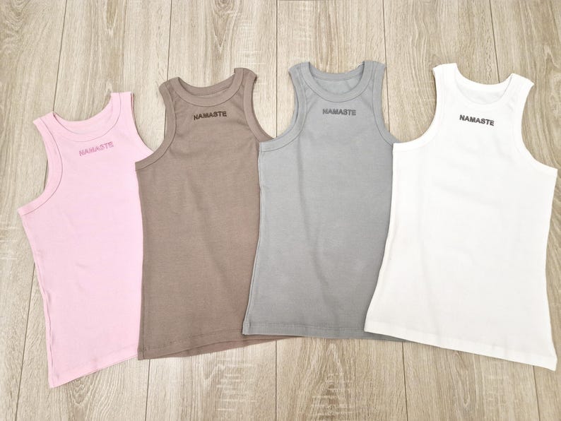K&ouml;nnte beinhalten: Vier &auml;rmellose Tanktops in verschiedenen Farben: rosa, braun, grau und wei&szlig;. Jedes Tanktop hat den Schriftzug "NAMASTE" auf der Vorderseite.