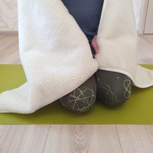 May include: A white fuzzy blanket draped over a person sitting on a green yoga mat. The person is wearing black leggings with a green abstract pattern.