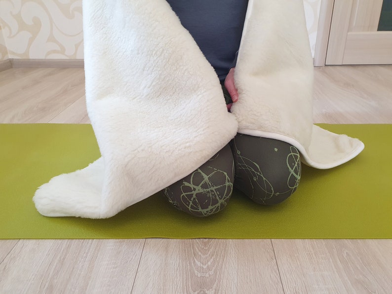May include: A white fuzzy blanket draped over a person sitting on a green yoga mat. The person is wearing black leggings with a green abstract pattern.