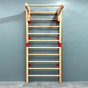 May include: A wooden wall-mounted stall bar with a horizontal top bar and multiple rungs. The wooden frame is light-colored, with red metal brackets securing the rungs. The stall bar is designed for exercise and stretching.